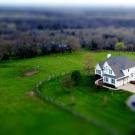 Aerial tilt-shift photograph of white farmhouse by a pond and winding driveway in green fields.